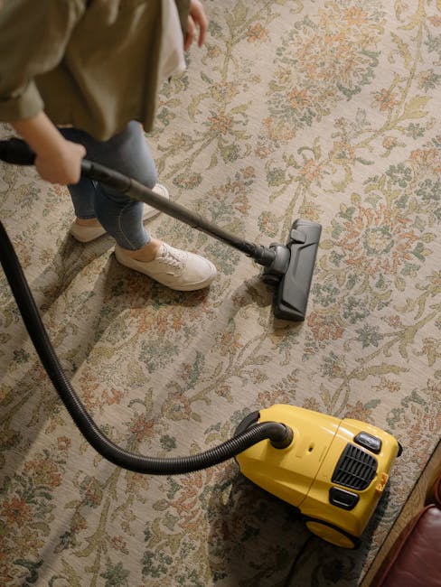 A person is using a vacuum cleaner with a yellow body and black hose to deep clean a large, patterned area rug in a residential room. The rug features intricate floral designs in soft pastel shades of